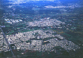 写真（当地区は、ＪＲ恵み野駅（写真中央左）周辺に位置し、団地の中央に帯状に恵み野中央公園が配置されている。）